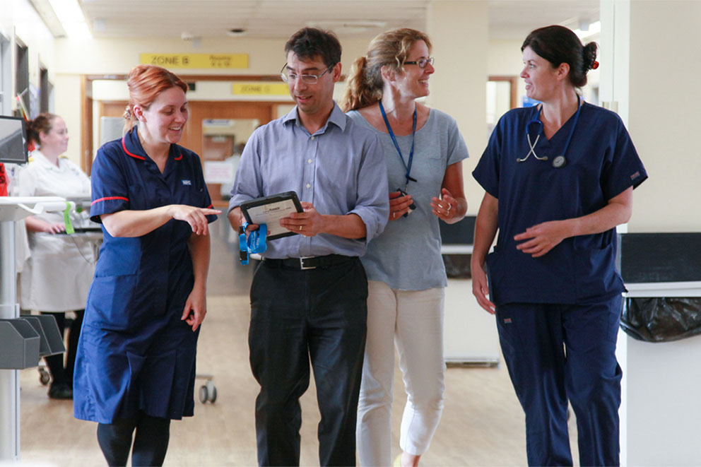 Picture with four clinical staff walking down the aisle of ward 30, using Wirral Millennium to discuss a patient treatment, Wirral University Teaching Hospital